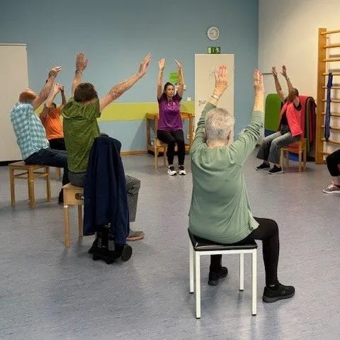 Teilnehmer führen Atemübungen im Sitzen durch, Hände hochgestreckt, in einem hellen Raum.