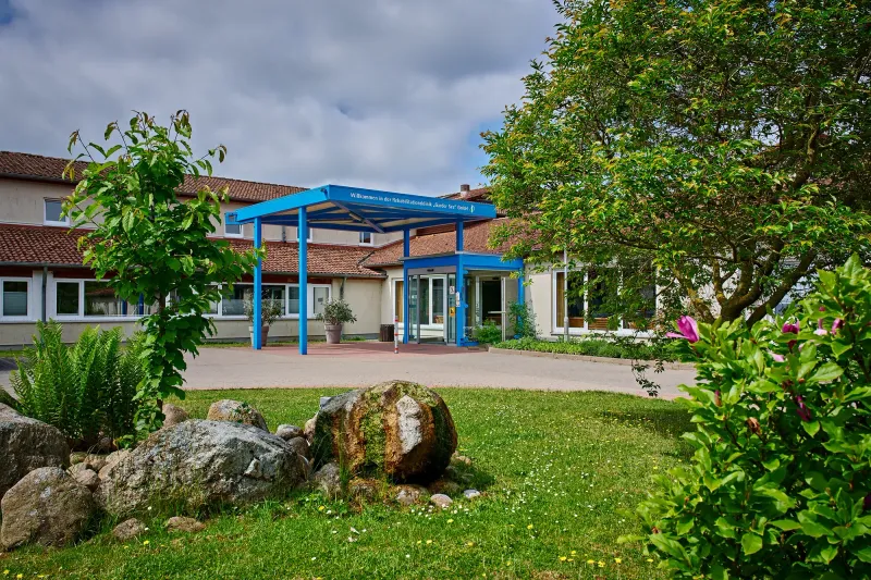 Eingang zur Rehabilitationsklinik mit blauer Überdachung und begrüntem Außenbereich.