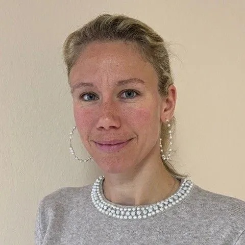 Frau mit blonden Haaren und Perlenohrringen lächelt vor neutralem Hintergrund.