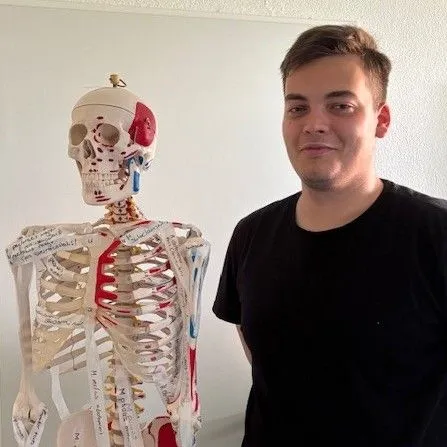 Junger Mann steht neben einem anatomischen Skelettmodell in einem hellen Raum.