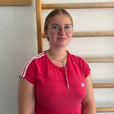 Junge Frau mit Brille und rotem T-Shirt steht vor einer Wand mit Holzregalen.