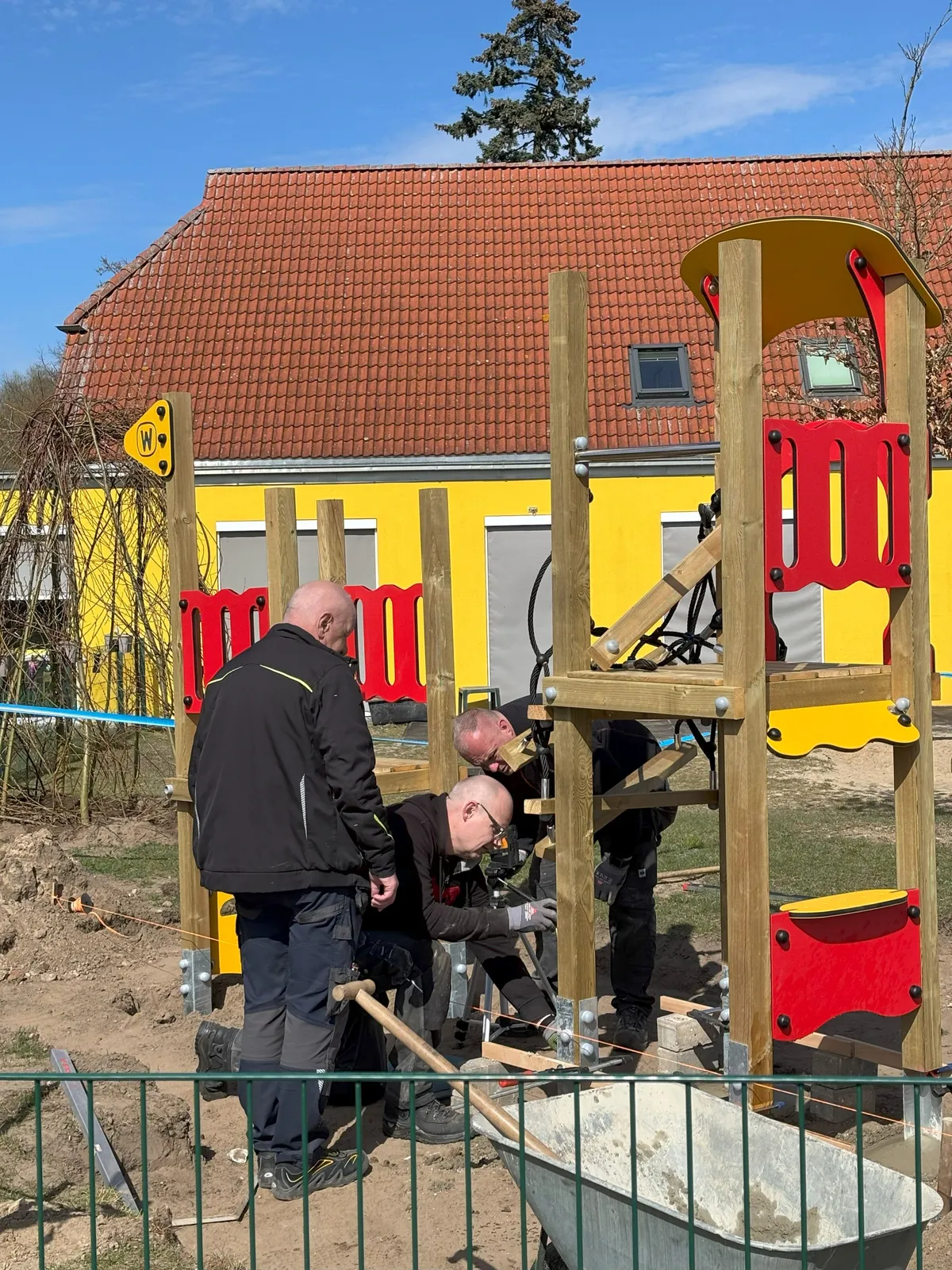 Drei Arbeiter montieren ein neues Spielgerät auf einem Spielplatz.