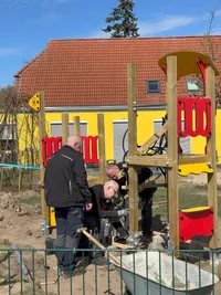 Drei Arbeiter montieren ein neues Spielgerät auf einem Spielplatz.