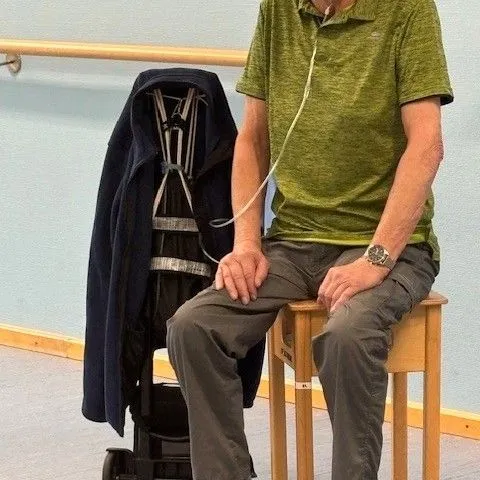 Mann sitzt auf einem Stuhl neben einem mobilen Sauerstoffgerät, trägt eine Sauerstoffmaske.