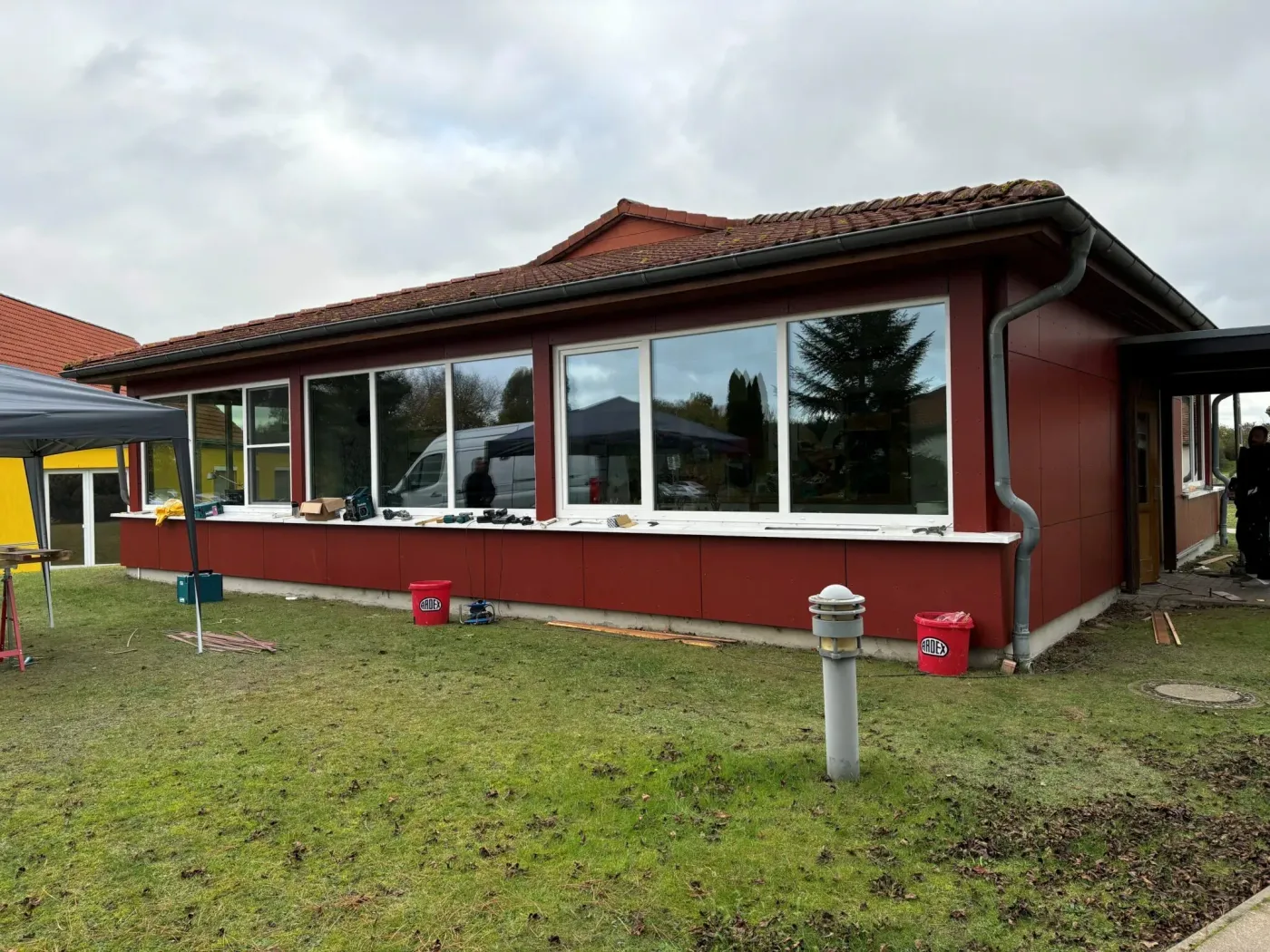 Roter Pavillon mit großen Fenstern, Baustellenutensilien und grünem Rasen im Vordergrund.
