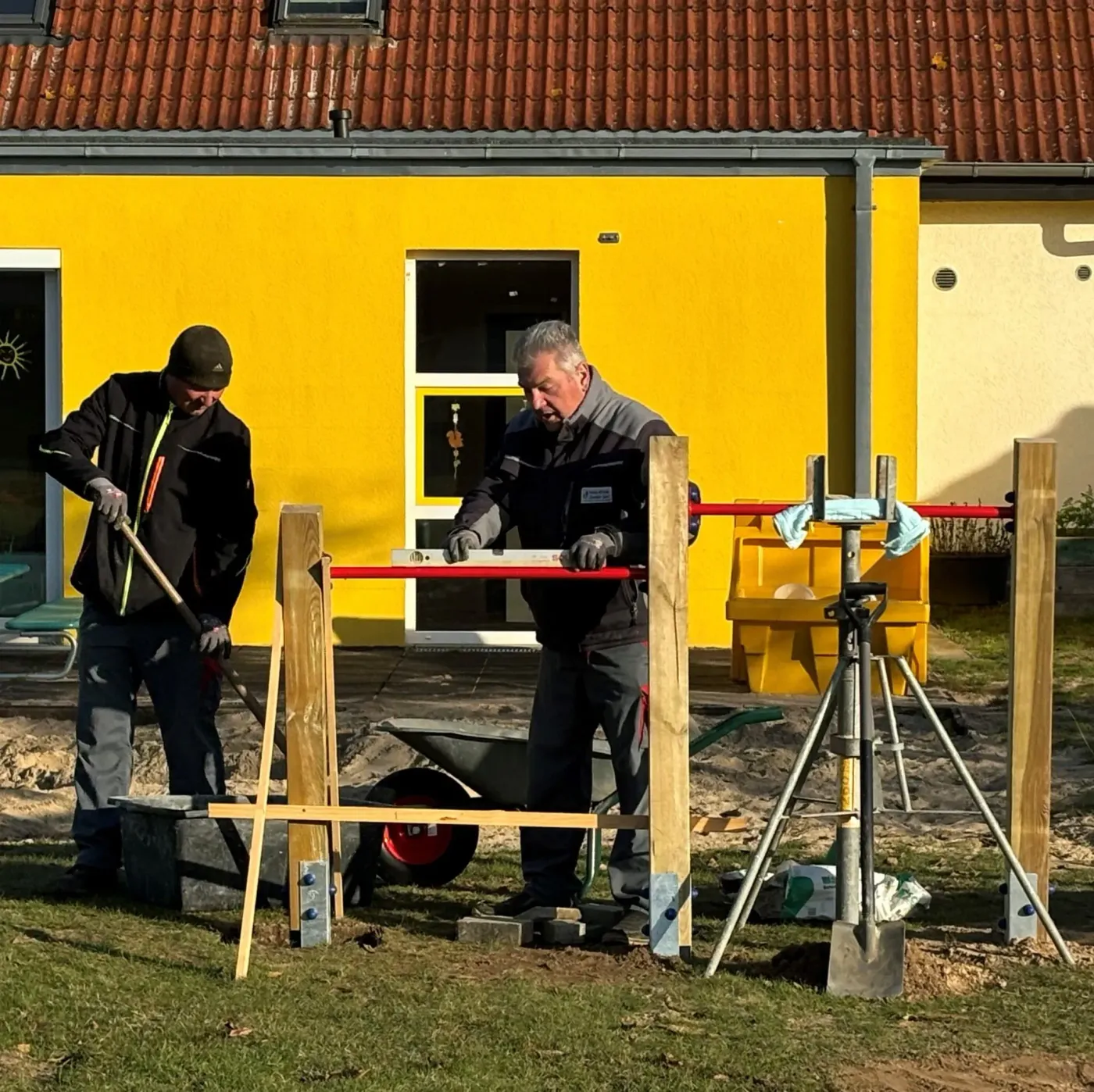 Zwei Arbeiter bauen einen Spielplatz, während sie Holzpfosten und Werkzeuge verwenden.